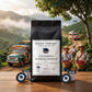 Colombian Supremo 100% Purace Coffee Pods Single-serve by Roast Theory. A black bag of Roast Theory Colombian Supremo Purace coffee stands on a wooden table, surrounded by two single-serve coffee pods and two round blue-and-white capsules. The background shows a scenic coffee plantation with workers carrying red coffee cherries, mountains, and vintage trucks.